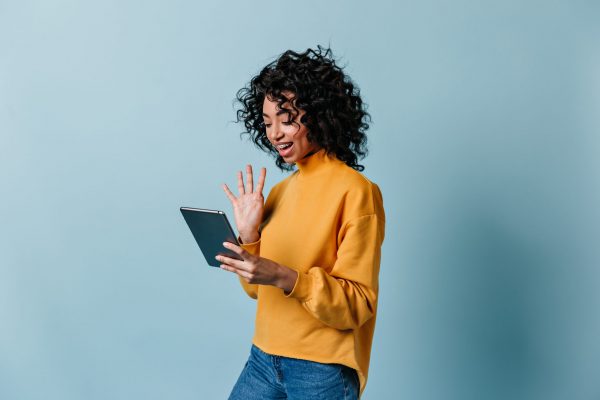 Smiling girl waving hand at digital tablet screen. Charming curly woman using video chat.